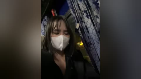 Media: A video of a woman with shoulder-length hair, wearing a white mask and black clothing, sitting in a dimly lit subway car with blue and white patterned curtains in the background.