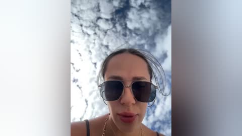 Media: Video of a woman with dark sunglasses, brown hair tied back, standing in front of a cloudy sky. She wears a gold necklace and a black top.