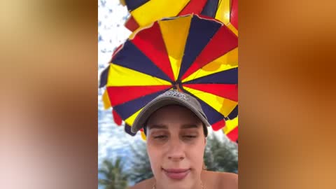 Media: Video of a young man with light skin and short brown hair wearing a colorful, umbrella-like headdress with red, yellow, and blue stripes. Background features blurred palm trees and a mountainous landscape under a bright, sunny sky.