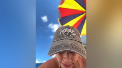 Media: Video of a smiling woman wearing a gray baseball cap with a white logo, in a bright, sunny beach scene with a vibrant, multicolored beach umbrella in the background.