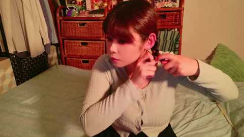 Media: Video of a young Asian woman with brown hair, wearing a light gray cardigan, adjusting a pink earring in a cozy bedroom with wooden furniture and a green bedspread.