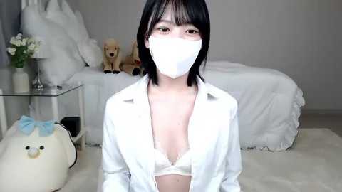 Media: Video of a young East Asian woman with short black hair, wearing a white face mask, a partially unbuttoned white shirt revealing a white bra, in a cozy bedroom with plush toys and a plush bed.