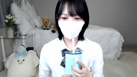 Media: Video of an East Asian woman with short black hair, wearing a white mask and white blouse, drinking blue milk from a clear glass jar, in a minimalist, white-themed bedroom with stuffed toys.