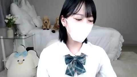 Media: Video of an East Asian girl with short black hair and light skin wearing a white mask, white sweater, and plaid bow tie, sitting on a plush white rug in a minimalist bedroom with white bedding and stuffed animals.