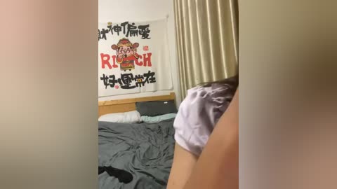 Media: Video of a woman in light pink pajamas, leaning over a bed with a gray blanket. Behind her, a white wall with a colorful \"Rokujouma no Isekai Shoukanshitsu\" poster.