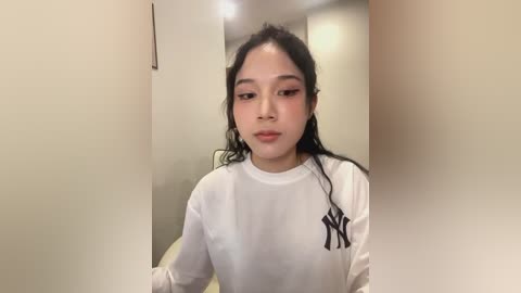 Media: Video of a young Asian woman with long, wavy black hair, wearing a white New York Yankees sweatshirt, standing in a narrow hallway with beige walls and a blurred background.