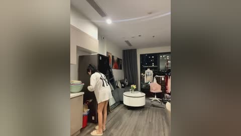 Media: Video of a modern apartment with a woman in a white robe and pink slippers sorting laundry. The room features a mix of contemporary and minimalist decor, including a white round coffee table and a gray sofa.