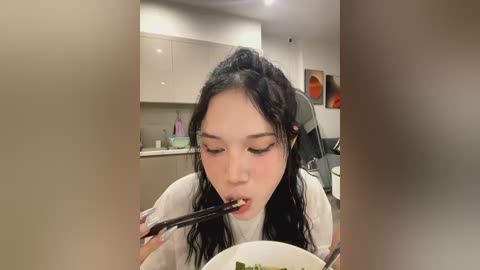 Media: Video of an Asian woman with long black hair, wearing a white t-shirt, eating noodles with chopsticks in a modern kitchen with beige cabinets and orange artwork.