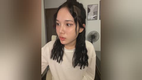 Media: A video of an East Asian woman with straight black hair in a white long-sleeved top, sitting in a dimly lit room with a fan and a framed abstract art on the wall.