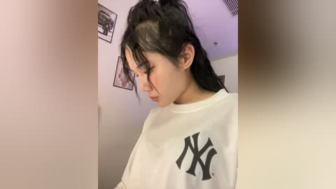 Media: Video of an Asian woman with long black hair in a high ponytail, wearing a white New York Yankees t-shirt, standing in a room with framed black-and-white photos on the wall.