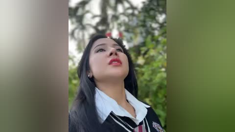 Media: Video of a woman with long black hair, wearing a black blazer with a white shirt and a red tie, standing in a lush, green outdoor setting.