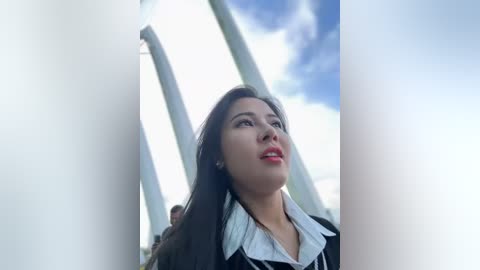 Media: Video of a young East Asian woman with long black hair and fair skin, wearing a white blouse and black jacket, standing against modern white arches under a partly cloudy sky.