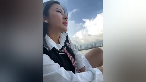 Media: Video of a young Asian woman in a black vest over a white shirt, with a red tie, sitting by a window overlooking a city skyline.