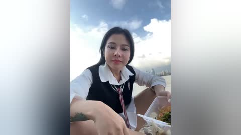 Media: Video of an Asian woman with straight black hair, wearing a white blouse and black vest, eating noodles on a bridge under a partly cloudy sky.