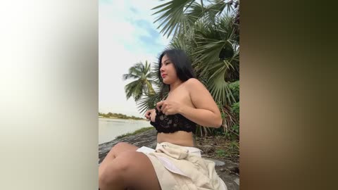 Media: Video of a young East Asian woman with long black hair, medium breast size, and medium skin tone, sitting on a rocky beach with palm trees, wearing a black lace bra and white skirt.