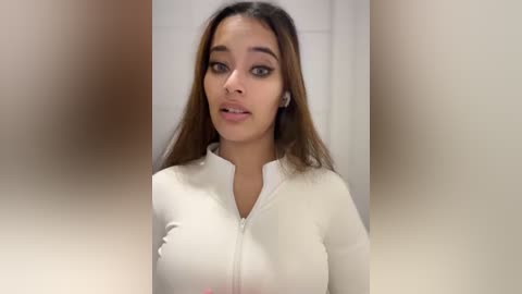 Media: Video of a young woman with medium skin tone and long, straight brown hair, wearing a white zip-up top. She has a neutral expression and is standing against a blurred beige background.