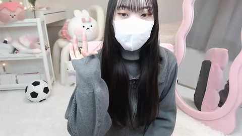 Media: Video of an East Asian woman with long black hair, wearing a white surgical mask, grey sweater, and jeans, in a pastel-themed room with plush toys, a soccer ball, and pink gaming chair.