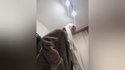 Media: A video captures a person, likely elderly, wearing a white robe and a brown blanket, bending over at the waist, appearing to be in pain. The setting is a dimly lit room with beige walls and a red curtain.