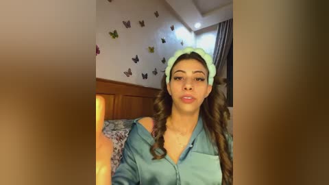 Media: Video of a young woman with long, wavy brown hair, wearing a light green cap and a teal blouse, sitting on a bed with a wooden headboard. The background features butterfly wall decals and a partially visible window.