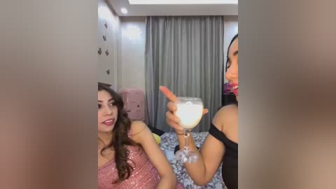 Media: Video of two women in a modern bedroom. One, with long brown hair, wears a pink sequin dress. The other, with short black hair, holds a drink and wears a black top.