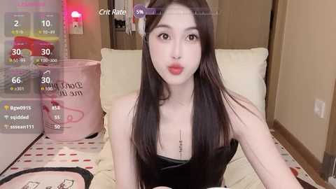 Media: A video of a young Asian woman with long black hair, fair skin, and red lipstick, wearing a black strapless top, sitting on a bed with a beige pillow, in a dimly lit room.