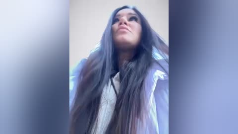 Media: Video of a woman with long, straight black hair, wearing a white blouse, standing against a blurred background. Her expression is contemplative.