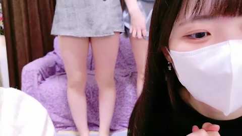 Media: Video of an Asian woman wearing a white face mask, black top, and grey shorts, standing in front of a purple armchair, with another person's legs visible behind her.