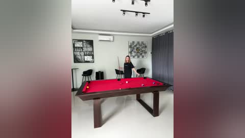 Media: A video of a modern, minimalist room with a red pool table, a woman in a black top, and black chairs against grey walls.