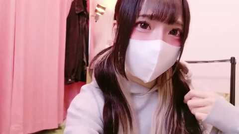 Media: A video of a young Asian woman with long, dark hair, wearing a white face mask and a light grey sweater. She stands in a pink-draped room with hanging clothes and a metal bed frame in the background.