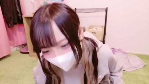 Media: Video of an Asian woman in a pink room with a white mask covering her mouth, leaning forward on green carpet, wearing a light grey jacket.