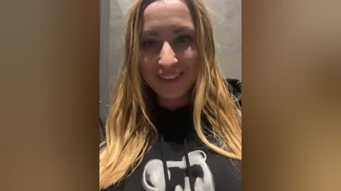 Media: Video of a smiling, light-skinned woman with long, wavy blonde hair, wearing a black t-shirt with a white skull design. She stands indoors against a plain, light-colored wall, her hair slightly disheveled.