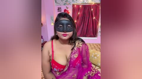 Media: Video of a woman with medium brown skin, wearing a red saree with gold embroidery, black mask, and red lipstick, in a pink room with floral wallpaper, fairy lights, and a red curtain.