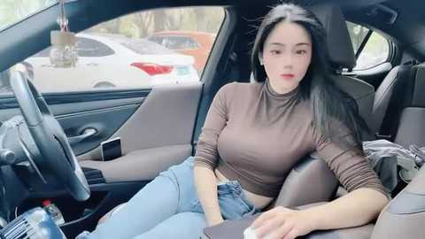 Media: Video of a young East Asian woman with long black hair, fair skin, and medium breasts, wearing a tight brown turtleneck and blue jeans, sitting in a car with a parked white SUV outside.