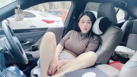 Media: Video of a young East Asian woman with long black hair, wearing a tight, light brown top, sitting in a modern car, legs spread, masturbating.