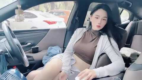 Media: Video of an East Asian woman with long black hair, fair skin, wearing a brown crop top and white cardigan, sitting in a car with a messy interior.