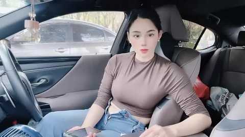 Media: Video of a young Asian woman with long black hair, wearing a brown crop top and blue jeans, sitting in a car seat, with a gray car in the background.