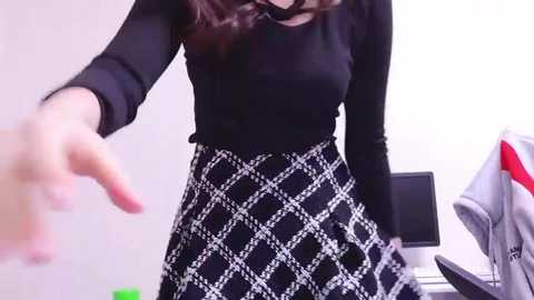 Media: Video of a woman with long brown hair, wearing a black top and a black and white diamond-patterned skirt, extending her hand, in a minimalistic office setting.