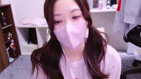 Media: Video of an Asian woman with long brown hair and light skin wearing a white face mask, white top, and sitting in a cluttered bedroom with a desk, shelves, and hanging clothes in the background.