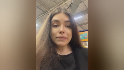 Media: Video of a young woman with long, straight brown hair, light skin, and dark eyes, wearing a black top, standing indoors in a warehouse with industrial ceiling and shelving.