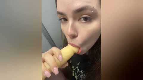 Media: Video of a young woman with fair skin, brown hair, and a nose piercing, licking a large, yellow dildo while wearing a black t-shirt with a graphic design.