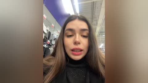 Media: Video of a young woman with long, straight, dark brown hair, wearing a black turtleneck sweater, standing in a brightly lit, modern clothing store with racks of clothes in the background.