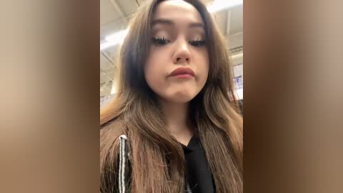 Media: A close-up video of a young woman with long, wavy brown hair, wearing a black top, looking serious with her lips slightly parted, taken indoors with fluorescent lighting.