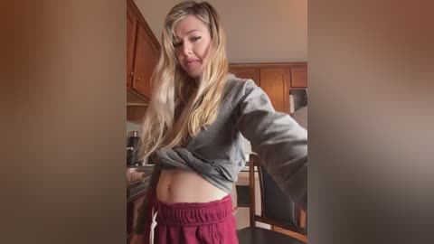 Media: A candid video of a pregnant woman in a kitchen, lifting her gray sweatshirt to reveal her bare stomach, wearing red pajama pants.