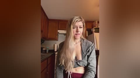 Media: A video of a blonde woman with long hair, wearing a grey hoodie, revealing her cleavage, standing in a dimly lit kitchen.