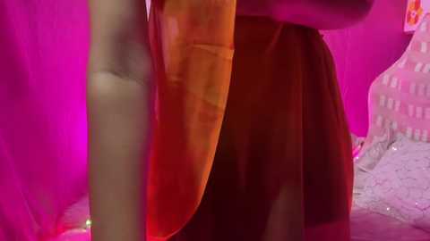 Media: Video of a person wearing a sheer, orange fabric, with vibrant pink and white background. The person's arm is visible, suggesting they are leaning against a wall.