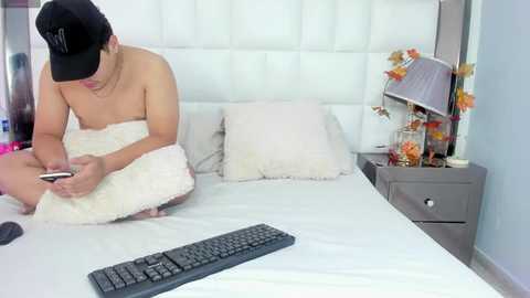 Media: Video of a shirtless man in a black cap, sitting on a white bed, texting on a smartphone. A black keyboard rests on the bed, and a gray nightstand with a lamp and decorative items is visible in the background.