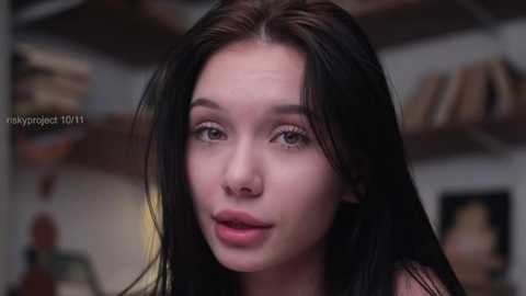 Media: Video of a young woman with fair skin, long black hair, green eyes, and full lips, wearing a sleeveless top. She is indoors, with blurred shelves in the background.