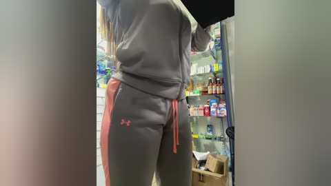 Media: Video of a person with long hair, wearing a grey sweatshirt and matching grey sweatpants with red Under Armour logo, standing in front of a fully stocked refrigerator.