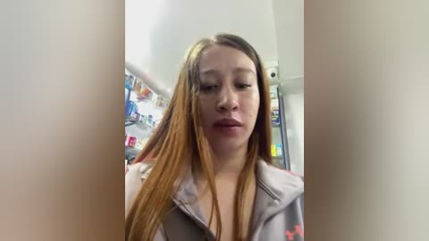 Media: Video of a young woman with straight, light brown hair, wearing a grey hoodie with red star accents, standing in a well-lit kitchen. Shelves filled with various items are visible in the background.