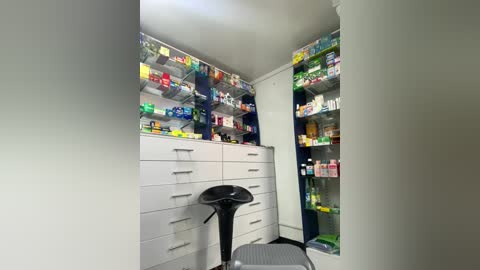 Media: A video of a well-lit, modern pharmacy with white walls, shelves filled with colorful medicines, a black stool, and a white cabinet.